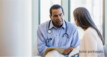 Actor portrayals of male doctor with stethoscope around his neck sitting with female patient while discussing documents